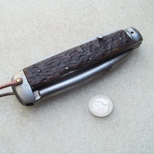 May include: A closed folding pocket knife with a textured dark brown handle, metal bolsters, and a copper-coloured bail loop at one end. A small silver-coloured coin is positioned next to the knife for scale, showing its relative size.