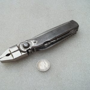 May include: A vintage silver-toned multi-tool featuring integrated pliers and a folding handle mechanism. The tool is marked with the word ELECTRIC and is positioned next to a small circular silver coin for scale, showing the tool's compact size.