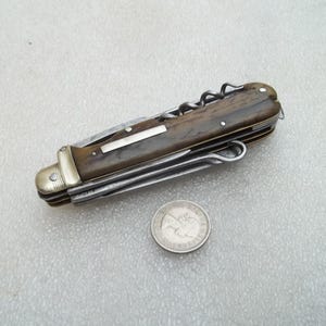 Può includere: Un coltellino svizzero multiuso con manico marrone testurizzato, una guardia in ottone e diversi utensili in acciaio ripiegati, tra cui un cavatappi. Una piccola moneta rotonda color argento è posizionata sotto il coltello chiuso.