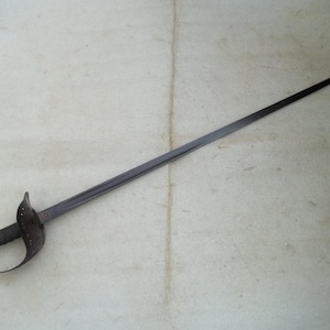 May include: A dark gray sword with a curved handguard and a long, pointed blade. The handle is wrapped in a textured material, and the guard has a decorative design. The sword is lying on a light-colored surface.