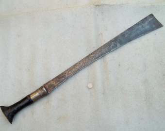 May include: A vintage machete with a long, slightly curved blade. The blade shows signs of wear and age. The handle is black with a silver-colored metal band near the blade. The machete is lying on a light-colored surface.