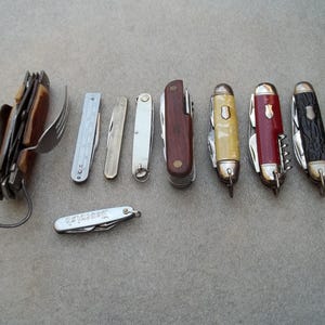 Op de afbeelding: Een verzameling van negen zakmessen met verschillende ontwerpen en functies. De messen hebben verschillende gekleurde handvatten, waaronder rood, bruin, zwart en wit. Sommige messen hebben een klein vorkje aan het handvat.