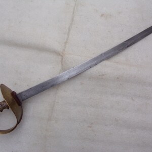 Op de afbeelding: Een zilverkleurig zwaard met een messing handvat en beschermkap. Het zwaard heeft een lange, rechte kling en een eenvoudig, afgerond handvat.