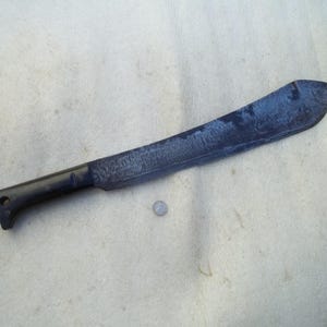 以下が含まれることがあります： 黒い柄と、わずかに錆びた暗い色の刃を持つ、大きく湾曲したマチェーテ。 刃は湾曲した形状で、鈍い仕上がりです。 マチェーテは明るい色の表面に置かれています。