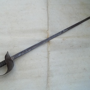 May include: A vintage, antique sword with a dark, weathered appearance. The blade is long and slender, with a pointed tip. The hilt features a curved guard and a textured grip. The overall design suggests a historical or collectible item.