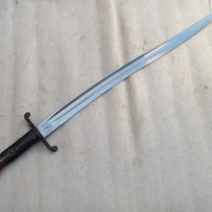 May include: A vintage bayonet featuring a long, silver blade and a dark wooden handle. The blade is straight and tapers to a point. The handle has a crossguard and a pommel. This bayonet is likely from a historical military collection.