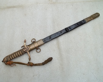 May include: An antique sword with a black blade and an ornate gold-coloured hilt. The hilt features a crossguard, rings, and decorative elements. A brown cord is attached to the hilt. The sword is likely a ceremonial or historical item.