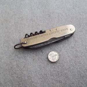 Puede incluir: Un cuchillo de bolsillo plegable de latón con un sacacorchos, una regla y una hoja. El cuchillo está grabado con las palabras "Pegley-Davies Anglers' Knife".