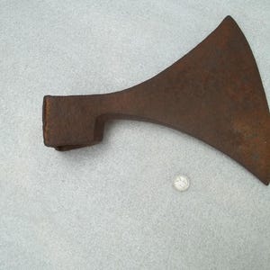 May include: A rusted iron axe head with a wide, curved blade and a rectangular socket for a handle. The metal surface shows significant oxidation and texture. A small, circular silver-colored coin is positioned below the axe head for scale.