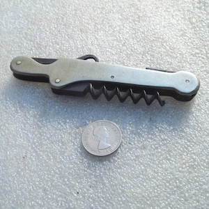 以下が含まれることがあります： 銀色の金属製ハンドルと黒いコルク抜きを備えた折りたたみ式のマルチツールポケットナイフ。ツールは、エリザベス2世の横顔が刻印された銀色のコインの隣に置かれており、サイズ比較の目安となっています。
