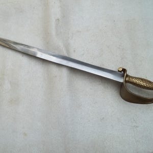 May include: A saber with a polished steel blade and a brass handle. The handle has a textured, fish-scale pattern and a curved guard. The sword is displayed on a light-colored surface.