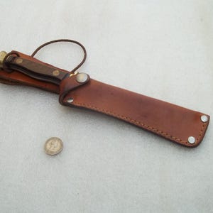 Puede incluir: Un cuchillo de hoja fija con mango de madera marrón oscuro y pomo de latón, guardado en una funda de cuero color canela con remaches metálicos y cierre a presión. Una pequeña moneda metálica circular se sitúa junto a la funda.