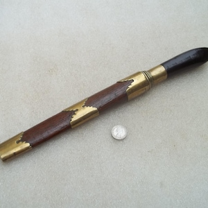 Pode incluir: Uma bainha de faca de madeira vintage com detalhes em latão. A bainha é marrom escuro com acessórios de latão nas extremidades e ao redor da alça. A alça é marrom mais escuro. Uma moeda está ao lado da bainha.