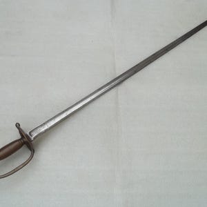 Puede incluir: Una espada de acero larga y delgada con una hoja puntiaguda y un canal central. La empuñadura consta de un mango de madera oscura y lisa, una guarda de metal sencilla y un guardamano curvo que conecta con el pomo en la base del mango.