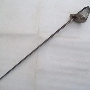 May include: A silver-colored sword with a decorative hilt. The hilt is pierced with holes and has a floral design. The sword is on a white background.