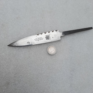 May include: A steel knife blade with a pointed tip and a serrated spine. The blade features an engraved crest and the text W. ROBB BALLATER & BRAEMAR BY APPOINTMENT. A small silver-colored coin is positioned below the blade for scale reference.