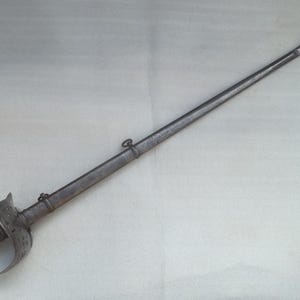May include: A metallic sword with a curved blade housed in a matching scabbard. The hilt features a prominent hand guard and a ribbed grip. Two small metal rings are attached to the scabbard, which terminates in a rounded metal tip.