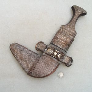 Puede incluir: Una daga antigua y ornamentada, con empuñadura y vaina de metal plateado. La empuñadura y la vaina están intrincadamente decoradas con patrones florales y geométricos. La daga está sobre una superficie clara, con una moneda como escala.
