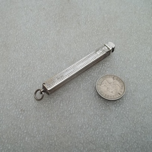 Może przedstawiać: Srebrzysty, sześciokątny, podłużny metalowy wisiorek z wygrawerowanymi oznaczeniami liczbowymi na całej długości, przypominający miniaturową linijkę. Do jednego końca przymocowane jest małe kółeczko, obok znajduje się okrągła srebrna moneta.