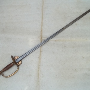 Puede incluir: Una espada antigua y larga con una hoja plateada y delgada. La empuñadura tiene un mango de madera, una guarda de latón y un pomo de latón. La espada se exhibe sobre una superficie clara.
