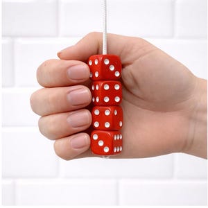 May include: Four red dice with white spots, strung on a white cord. The dice are held in a hand against a white tiled background. Each die shows a different number of spots. The dice are made of a hard material.