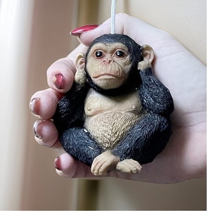 Op de afbeelding: Een klein zwart en bruin chimpanseefiguur met zijn handen over zijn oren. Het figuur hangt aan een wit koord en wordt vastgehouden door een hand.