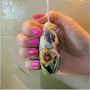 Può includere: Tirante per la luce in ceramica ovale con un motivo floreale. Presenta girasoli, foglie e altri fiori nei toni del giallo, rosa e blu. Attaccato a un cordino bianco.