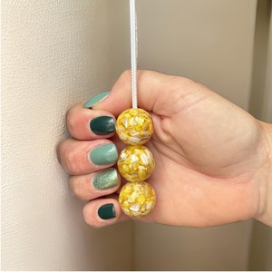 May include: Three yellow and white speckled round beads on a white cord. The beads are strung together and are being held by a hand.