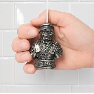 May include: A silver-colored Roman soldier bust toilet pull with the word "ROMAN" at the base. The pull is held by a hand against a white tile background. The pull is attached to a white cord.