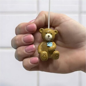 Peut inclure: Tirette de lumière en forme d'ours en peluche marron avec une fleur bleue. Suspendu à un cordon blanc, cet accessoire décoratif est idéal pour une chambre d'enfant ou une crèche.