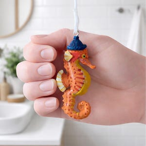 Pode incluir: Enfeite de cavalo-marinho, predominantemente laranja com detalhes amarelos e vermelhos, suspenso por um cordão branco. O cavalo-marinho usa um pequeno chapéu azul. Ideal para decoração com tema náutico.