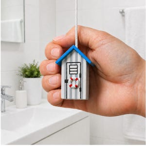 May include: A miniature beach hut light pull with a blue roof and a white and grey striped body. The hut has a small door and a red and white life preserver. The pull is suspended by a white cord, ideal for a bathroom.