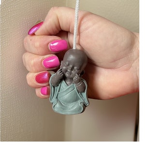 May include: A small, brown, smiling Buddha figurine with closed eyes, wearing a light blue robe, is attached to a white cord. The figurine is being held by a hand with pink nail polish.