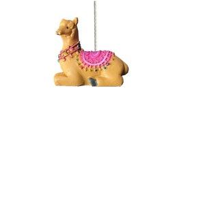 May include: A tan llama ornament with a pink and multi-coloured saddle. The llama is lying down with its head turned to the side.