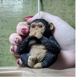 Op de afbeelding: Een klein zwart en bruin aapje figuur met zijn handen voor zijn ogen. Het figuur wordt vastgehouden in een hand met rode nagellak.