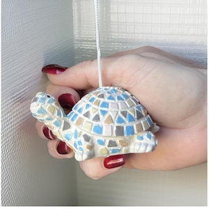 May include: A mosaic turtle ornament with a white pull cord. The shell is made of small tiles in blue, beige, and brown. The figurine is held in a hand with red nail varnish.