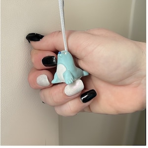 Könnte beinhalten: Eine hellblaue Robbenfigur mit einer weißen Schnur. Die Robbe hält ein kleines weißes Objekt im Maul. Die Figur wird von einer Hand mit schwarzem und weißem Nagellack gehalten.