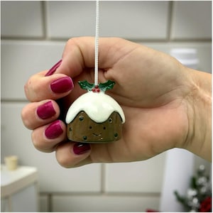 May include: A miniature Christmas pudding decoration, brown with white icing and a holly sprig. The ornament has a smiling face and is held by a white string. A festive Christmas tree ornament.