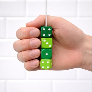 May include: A hand holds a string of four green dice. The dice are in varying shades of green and have white dots representing numbers. The background is a white tiled wall.