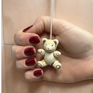 May include: A small, off-white teddy bear light pull with a cream-colored body and brown accents. The bear is suspended by a white cord. The image shows the bear held in a hand with dark red nail polish.