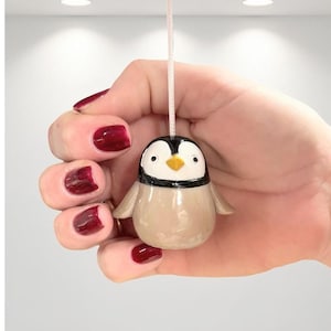 Pode incluir: Um pequeno ornamento de pinguim de cerâmica com cabeça preta e branca, bico amarelo e corpo bege. O pinguim está suspenso por um fio fino esbranquiçado. Um item decorativo fofo.