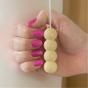 May include: A light beige, four-sphere pull chain with a white cord, held by a hand with bright pink nail polish. The pull chain is likely for a light fixture or window covering.