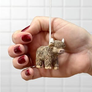 May include: A miniature Highland cow figurine with a shaggy brown coat, small white horns, and a pink nose. The cow is suspended by a white string. The figurine is held in a hand. The background is a white tiled wall.