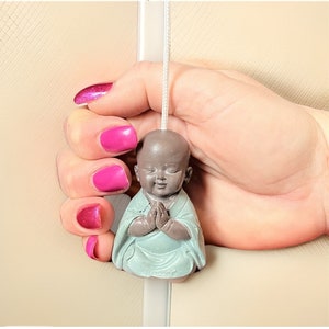 May include: A small, gray ceramic figurine of a meditating Buddha with a light blue robe. The figurine is attached to a white cord.