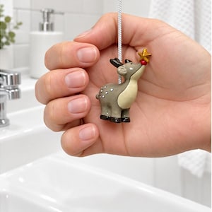 May include: A light pull featuring a small, grey deer figurine with white spots, dark hooves, and antlers. The deer is reaching upwards with its nose to touch a small, five-pointed gold star. The figurine is attached to a white braided cord.