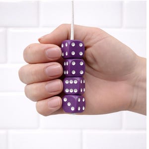 May include: A stack of four purple dice with white dots, strung together on a white cord. The dice are held in a hand against a white tile background. Each die has a different number of dots.