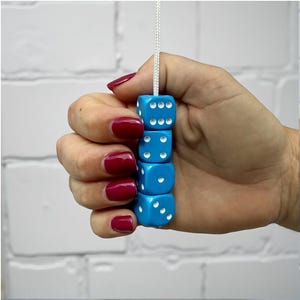May include: A stack of five turquoise dice, each with white dots, strung together on a white cord. The dice are held in a hand with red nail polish against a white brick background. The dice are a fun game accessory.