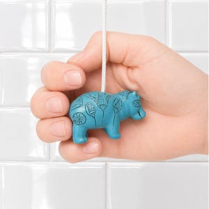 Puede incluir: Un tirador de luz turquesa con forma de hipopótamo con detalles grabados decorativos, suspendido por un cordón blanco. El hipopótamo se sostiene sobre un fondo de azulejos blancos.
