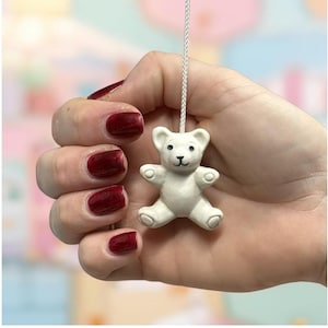 May include: A small, white teddy bear light pull with a black nose and eyes, suspended by a white cord. The bear is held in a hand with red nail polish, against a soft, colorful background.