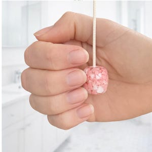 以下が含まれることがあります： 正方形のピンクと白の装飾的なライトプル。プルは、ピンクと白の小さな不規則な破片でできており、クリーム色のコードに通されています。背景はぼやけたバスルームの設定です。